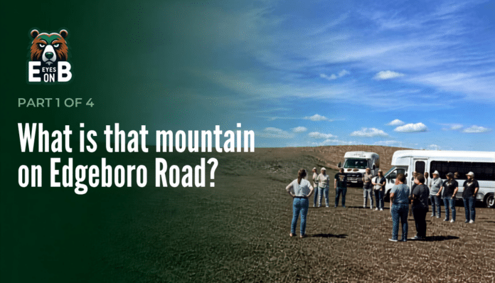 A group of about a dozen visitors stands on bare graded ground at the Edgeboro Road landfill beside two white shuttle buses, under a blue sky with scattered clouds. Overlaid title card reads: Eyes on EB, Part 1 of 4, What is that mountain on Edgeboro Road?