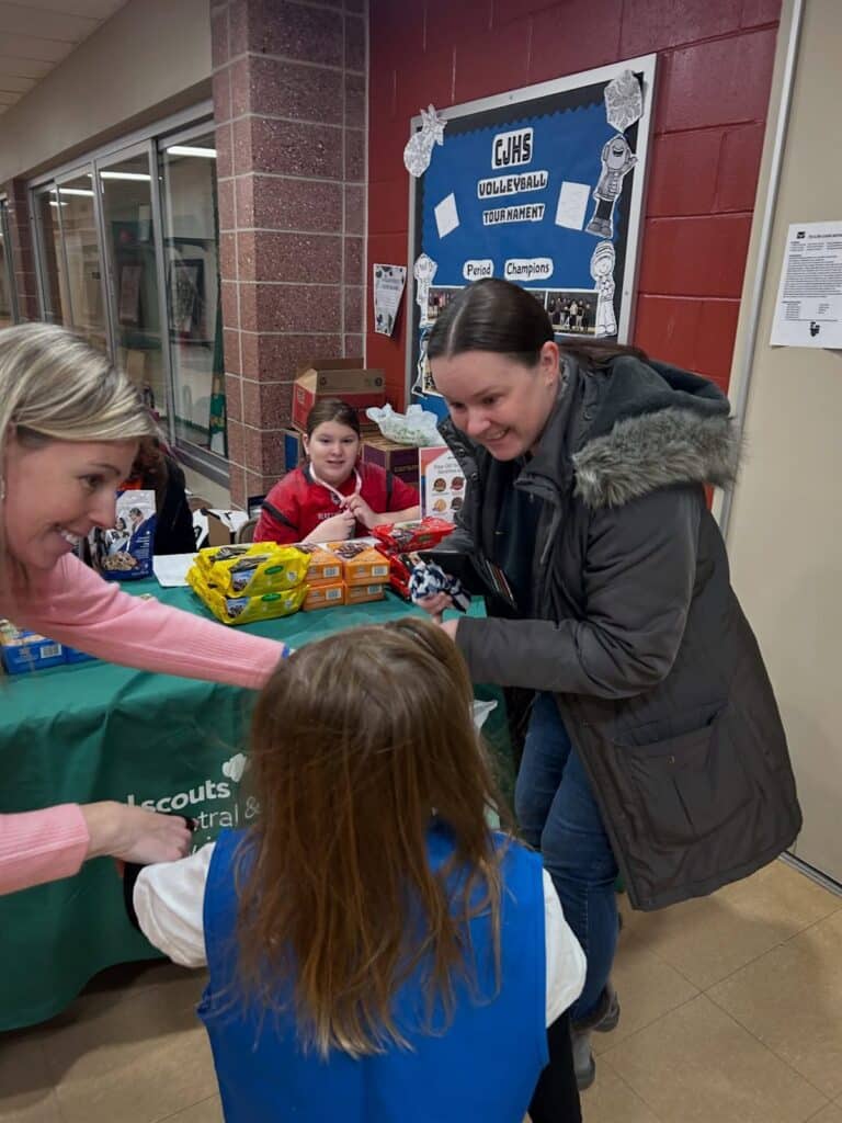A young Girl Scout in a blue vest, seen from behind, stands at a table stacked with boxes of Girl Scout cookies while two women lean toward her and another girl sits behind the table.