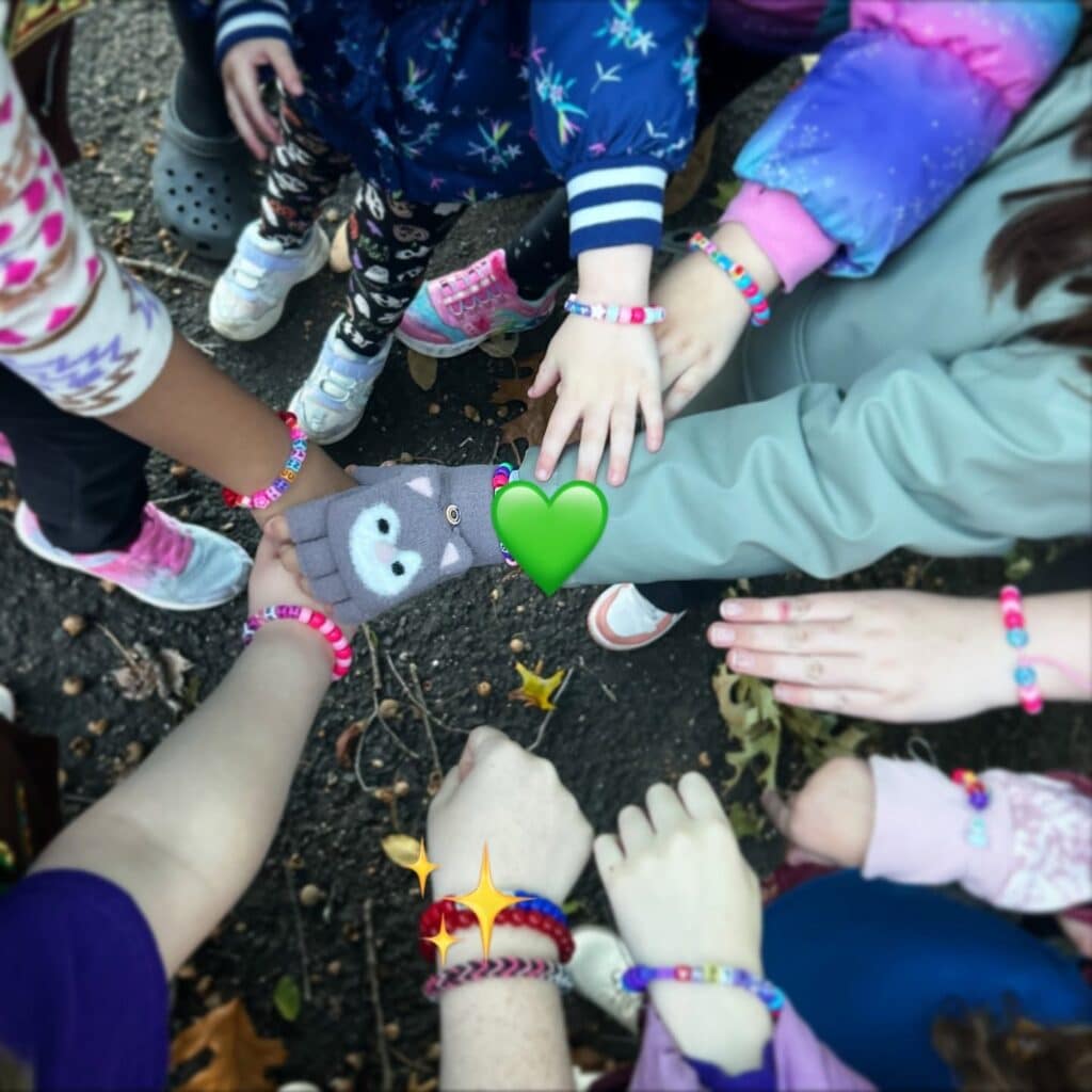 Seen from above, a circle of hands reaches toward the center over pavement and fallen leaves, many wearing colorful beaded bracelets.