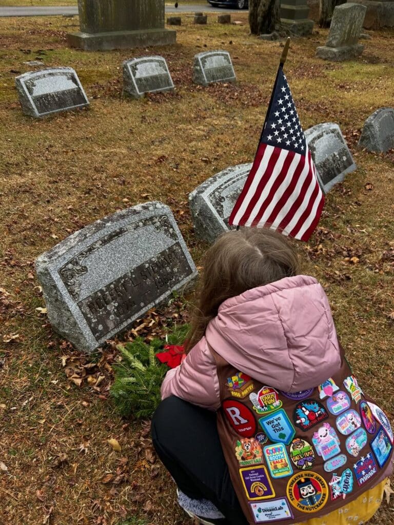 A girl in a pink jacket and a patch-covered Girl Scout vest, seen from behind, crouches beside a gray headstone where a wreath with a red bow and a small American flag have been placed.